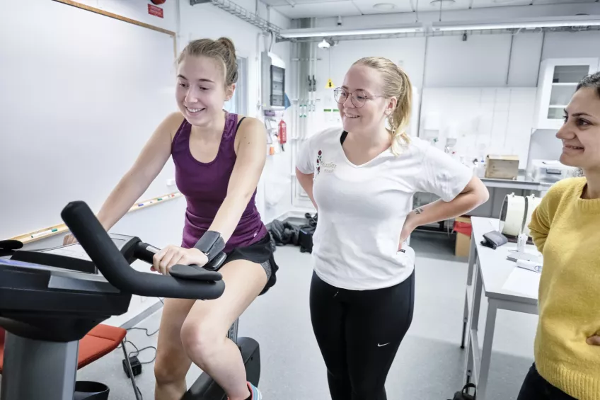 Three young women. One is on an exercise bike.